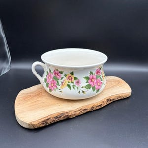 May include: A white ceramic chamber pot featuring a floral pattern of pink and yellow flowers with green leaves. The vessel has a single curved handle on the left side and a wide, rounded rim. The piece is displayed on a natural wood slab.