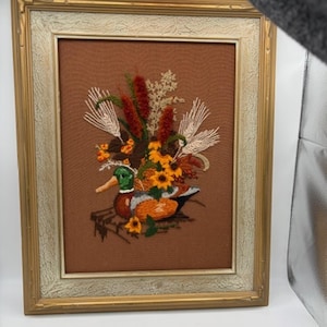 May include: Framed embroidery art featuring a duck with colorful plumage, surrounded by flowers and foliage. The artwork is set against a brown background and framed in a gold-colored frame with a cream-colored inner border. The embroidery is detailed.