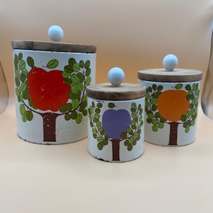 Könnte beinhalten: Set aus drei weißen Keramikbehältern mit Holzdeckeln und bunten Obstbaumdesigns. Die Behälter zeigen jeweils eine rote, orange und violette Frucht.
