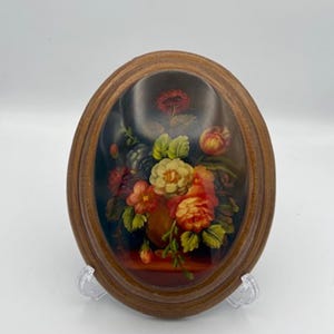 May include: Oval-shaped floral artwork in a wooden frame. The painting features a bouquet of red, yellow, and orange flowers in a vase. The artwork is displayed on a clear plastic stand against a white background.