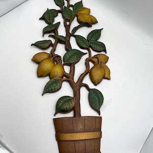 Könnte beinhalten: Eine Wanddekoration aus Metall, die einen Zitronenbaum mit grünen Blättern und gelben Zitronen darstellt. Der Baum steht in einem braunen Holztopf mit goldenen Akzenten.