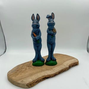 Può includere: Due statuine di coniglio in ceramica blu, una con una carota e l'altra con un fiore, in piedi su una superficie di legno.