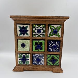 Puede incluir: Un pequeño mueble de madera con nueve cajones cuadrados. Cada frente de cajón presenta un diseño único de azulejo de cerámica colorido con patrones florales y un pomo azul central. El mueble tiene un acabado de madera oscura.