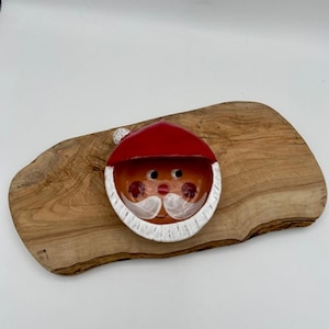Puede incluir: Una cara de Papá Noel de cerámica con un gorro rojo, barba blanca y mejillas rosadas. La cara de Papá Noel está colocada sobre una tabla de servir ovalada de madera. La tabla tiene un patrón de vetas de madera natural.