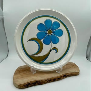 Peut inclure: Une assiette en céramique blanche avec un motif de fleur stylisé bleu et vert. L'assiette est sur un support en bois.