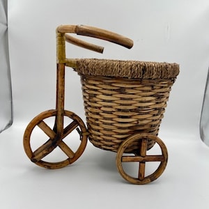 Pode incluir: Uma cesta decorativa em miniatura de vime em forma de bicicleta. A cesta é tecida com uma cor castanha natural e apresenta duas rodas de madeira e uma pega. A cesta foi concebida para fins decorativos.