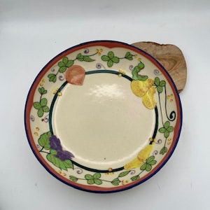 Op de afbeelding: Een decoratieve keramische plaat met een crèmekleurig midden en een kleurrijke rand. De rand heeft een rode buitenring, een blauwe binnenring en is versierd met groene klavers, gele peren en paarse druiven. De plaat staat op een houten oppervlak.