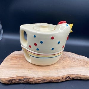 May include: Cream-colored ceramic teapot with a chicken design. The teapot features a red comb, yellow beak, and blue and red polka dots. It has a handle and a lid, and is decorated with horizontal stripes. The teapot is placed on a wooden cutting board.