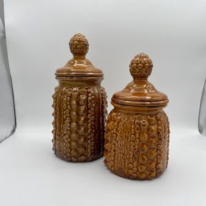 Op de afbeelding: Twee keramische potten met een bruine en gouden gespikkelde glazuur. De potten hebben een getextureerd, hobbelig oppervlak en een rond deksel met een knop.
