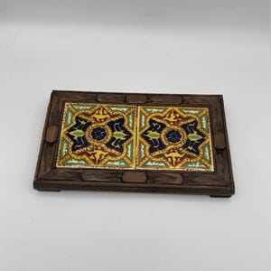 Puede incluir: Una bandeja decorativa de madera con dos baldosas de cerámica cuadradas. Las baldosas presentan un patrón floral repetitivo en azul, amarillo, verde y marrón. La bandeja es de madera marrón oscuro y tiene forma rectangular.