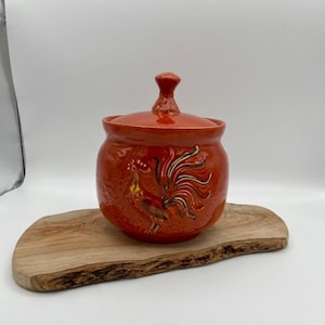 Può includere: Vaso in ceramica arancione con un design a gallo. Il vaso ha un coperchio e una superficie testurizzata.