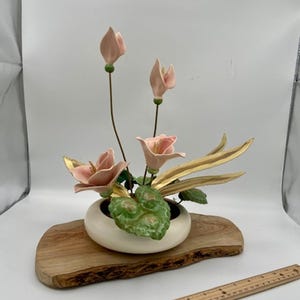 Może przedstawiać: Ceramiczna kompozycja kwiatowa z różowymi kwiatami i zielonymi liśćmi w białym wazonie. Kompozycja stoi na drewnianej podstawie. Po prawej stronie znajduje się linijka. Linijka ma około 30 cm długości.