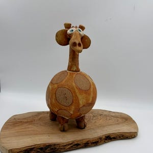May include: A whimsical ceramic giraffe sculpture with a round body and long neck, featuring brown spots on a tan background. The giraffe has blue eyes and is standing on a wooden base. A unique decorative item.