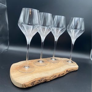 May include: Four clear wine glasses with a unique, angular design, resting on a natural, live-edge wooden serving board. The glasses have long stems and are arranged in a row, showcasing their elegant shape.