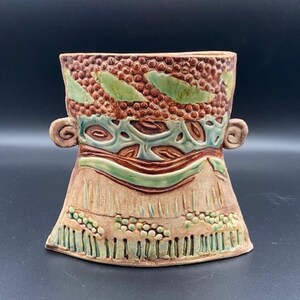 May include: A ceramic vase with a brown, green, and white design. The vase is shaped like a face with two small ears on the sides.
