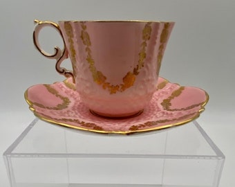 May include: A pink teacup and saucer with gold trim and floral designs. The cup and saucer are made of fine china and have a delicate, feminine look.