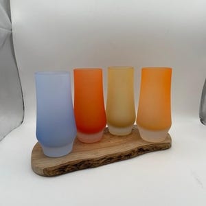 May include: Four frosted glass shot glasses in shades of blue, orange, yellow, and peach, arranged on a wooden serving tray.