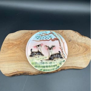 May include: A ceramic decorative plate featuring two deer resting under a tree. The plate has a circular shape with a pastel pink and blue background. The deer are black and white with detailed features, and the grass is green.