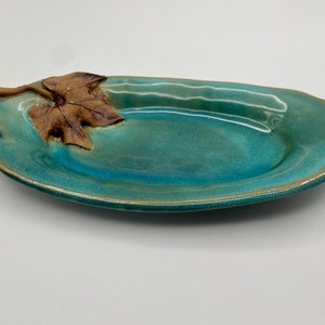 Op de afbeelding: Een teal keramische serveerschaal met een gesculpteerd blad en wijnstok handvat.