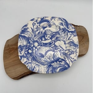 May include: A square, off-white plate with a blue cherub and floral design. The plate is placed on a wooden cutting board. The plate's design features a cherub, swirls, and floral elements.
