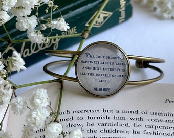 May include: A gold-toned bangle bracelet with a round charm that reads "The true secret of happiness lies in taking a genuine interest in all the details of daily life."  The charm is surrounded by a white background with black text. The bracelet is resting on a book with green cover and white text.