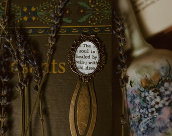 Könnte beinhalten: Ein goldenes Lesezeichen mit einem weißen ovalen Anhänger mit dem Text "The way a soul is healed by being with children".