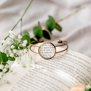 Puede incluir: Un brazalete de oro con un dije de vidrio que presenta una cita de Orgullo y Prejuicio: "Debes permitirme decirte cuán ardientemente te amo." El brazalete está sobre un libro abierto con flores blancas.