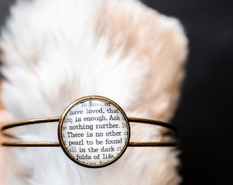 May include: A gold bangle bracelet with a round glass charm. The charm contains a printed page with the text "To love or have loved, that is enough. Ask for nothing further.  There is no other of pearl to be found... folds of life."