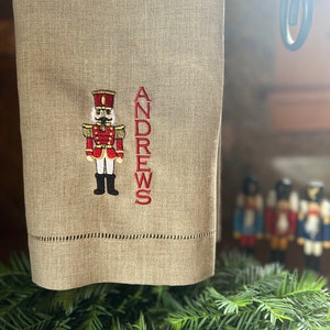 May include: A tan linen towel with a decorative embroidered nutcracker and the name "ANDREWS" in red. The nutcracker is in a red and gold uniform. The towel is hanging on a black metal bar, with greenery in the foreground.