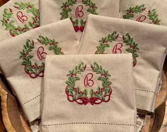 Puede incluir: Servilletas de lino beige, cada una bordada con una corona verde, bayas rojas y una letra "B" roja. Las servilletas están dispuestas en un cuenco de madera, con velas en el fondo. El bordado presenta un diseño festivo navideño.