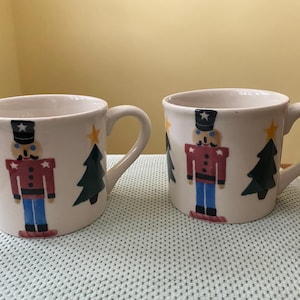 Peut inclure: Deux tasses en céramique blanche sur le thème de Noël. Chaque tasse présente un soldat casse-noisette peint, un sapin de Noël vert et une étoile jaune. Les tasses ont une anse incurvée et sont posées sur un fond clair.