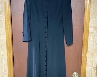 May include: A long, black dress with a high collar and long sleeves. The dress features a row of buttons down the front and is made of a smooth, flowing fabric. The dress is hanging on a hanger against a wooden door.