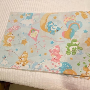 Könnte beinhalten: Ein hellblaues Care Bears-Flachbettlaken mit einem sich wiederholenden Muster aus Care Bears-Charakteren, Wolken, Sternen und Luftballons. Das Laken enthält den Satz "Snuggle up with sweet dreams!" und besteht aus einem weichen, leichten Stoff.