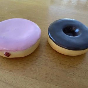 May include: Two round donut-shaped objects. One is a solid light pink glazed donut with a small red dot on the side. The other is a donut with a dark brown chocolate glaze and a central hole. Both have a light tan base color.