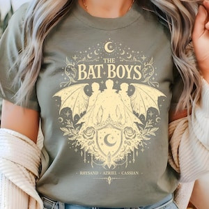 以下が含まれることがあります： 「The Bat Boys」の文字と、コウモリの羽を持つ3人の人物、三日月、花の要素が描かれたオリーブグリーンのTシャツ。Rhysand、Azriel、Cassianの名前がグラフィックの下に印刷されています。