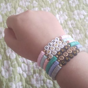 May include: A stack of colorful beaded bracelets with names spelled out in black and gold letters. The names are Remy, Mama, Elsie, Max, Sage, and Dad.