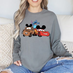 May include: A gray long-sleeve shirt with a graphic of Stitch, Mater, and Lightning McQueen from Disney's Cars and Lilo & Stitch. The shirt has a crew neck and long sleeves.