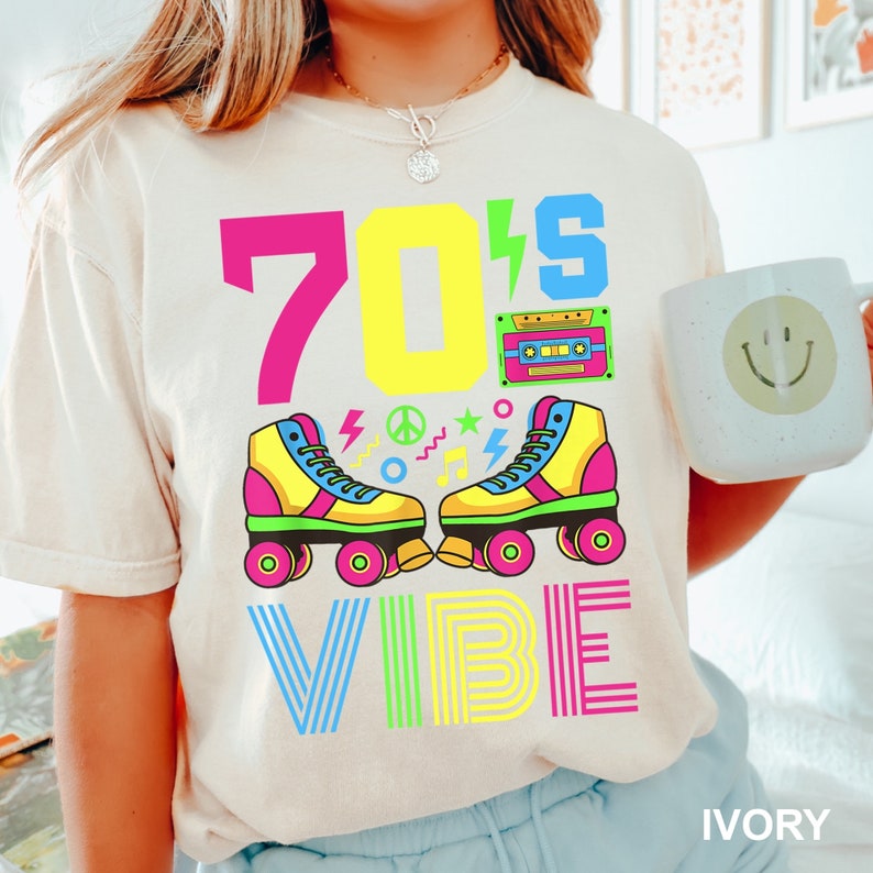 May include: A white t-shirt with a colorful graphic design featuring the text "70's VIBE" and a pair of roller skates. The graphic also includes a cassette tape and other retro-inspired designs.