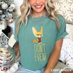 May include: Light green t-shirt featuring a cartoon fennec fox with a stern expression, wearing a red shirt. The text "DON'T EVEN!" is printed below the fox in yellow. The shirt is a light green color.