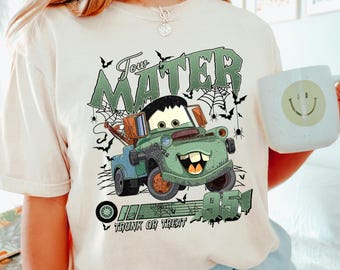 May include: Ivory-colored t-shirt featuring a cartoon image of Tow Mater from the movie Cars, with a Halloween theme. The design includes the text "Tow Mater", "95", and "Trunk or Treat", along with spiderwebs and bats. A white mug with a smiley face is also visible.