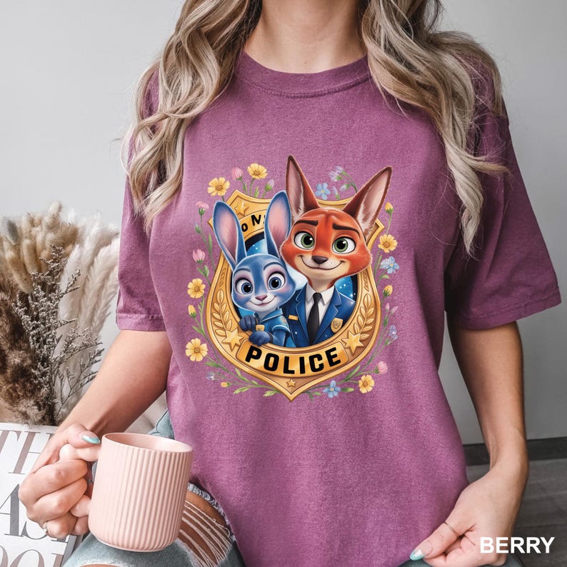 K&ouml;nnte beinhalten: Beerenfarbenes T-Shirt mit einem Cartoon-Abzeichen mit Judy Hopps und Nick Wilde aus Zootopia. Das Abzeichen tr&auml;gt die Aufschrift "POLICE" und ist von Blumen umgeben. Das Shirt ist ein kurz&auml;rmeliges Rundhals-Shirt.