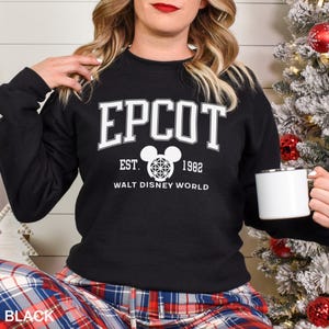 K&ouml;nnte beinhalten: Schwarzes Sweatshirt mit wei&szlig;em Text, der "EPCOT EST. 1982 WALT DISNEY WORLD" lautet. Das Design beinhaltet einen Mickey Mouse-Kopf mit einem geometrischen Muster. Das Wort "BLACK" befindet sich am unteren Bildrand.