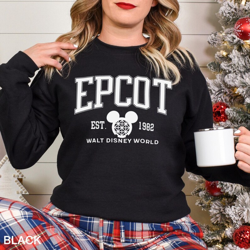 K&ouml;nnte beinhalten: Schwarzes Sweatshirt mit wei&szlig;em Text, der "EPCOT EST. 1982 WALT DISNEY WORLD" lautet. Das Design beinhaltet einen Mickey Mouse-Kopf mit einem geometrischen Muster. Das Wort "BLACK" befindet sich am unteren Bildrand.