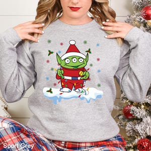 Puede incluir: Sudadera gris con un dise&ntilde;o festivo de un extraterrestre de dibujos animados. El extraterrestre lleva un gorro de Pap&aacute; Noel y un traje rojo, adornado con luces navide&ntilde;as, y est&aacute; sobre una base nevada. El dise&ntilde;o incluye copos de nieve y acebo.