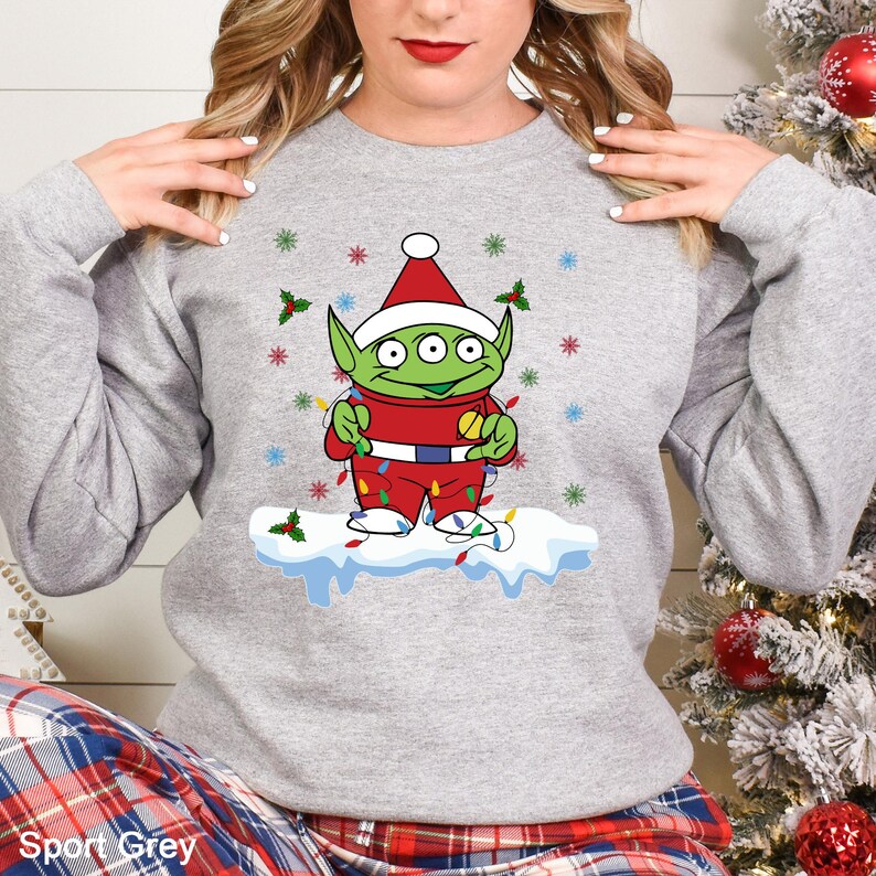 Puede incluir: Sudadera gris con un dise&ntilde;o festivo de un extraterrestre de dibujos animados. El extraterrestre lleva un gorro de Pap&aacute; Noel y un traje rojo, adornado con luces navide&ntilde;as, y est&aacute; sobre una base nevada. El dise&ntilde;o incluye copos de nieve y acebo.