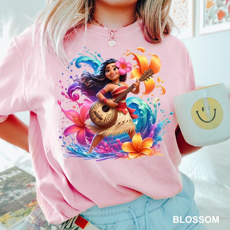 Puede incluir: Camiseta rosa con un personaje de dibujos animados tocando el ukelele, rodeado de flores coloridas y salpicaduras de agua. El personaje lleva un top rojo y blanco y una falda de hierba. Se sostiene una taza blanca con una carita sonriente amarilla.