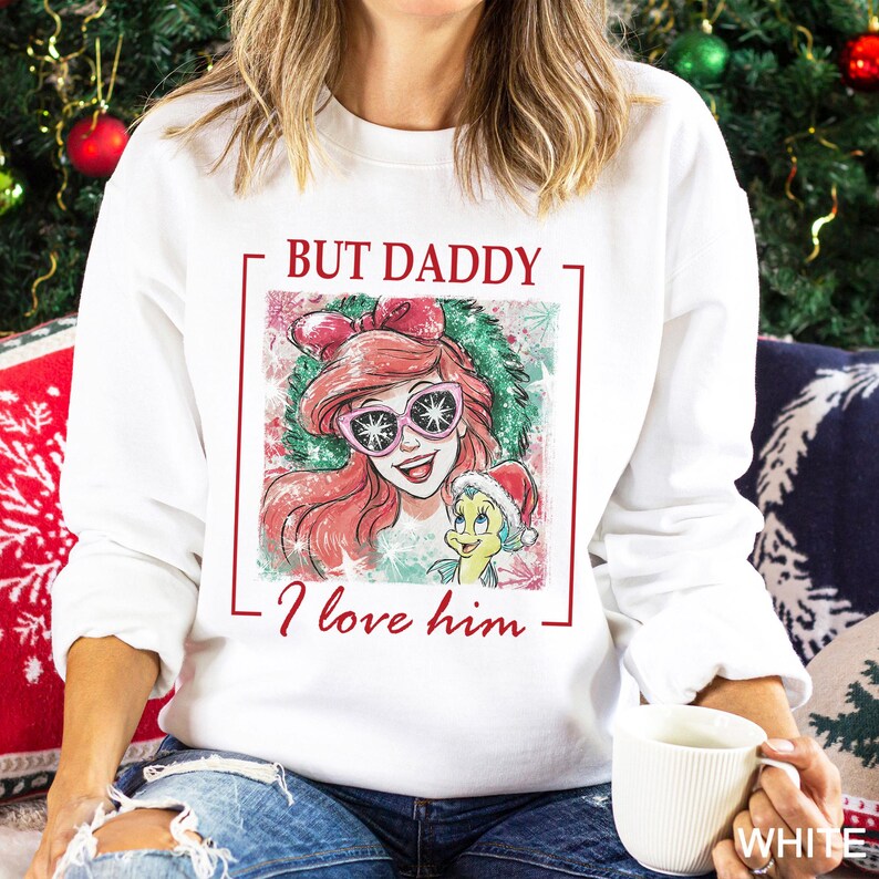 May include: White sweatshirt featuring a graphic of a cartoon character with red hair and sunglasses, and a yellow fish wearing a Santa hat. The text "BUT DADDY I love him" is printed in red. The sweatshirt is a festive holiday design.
