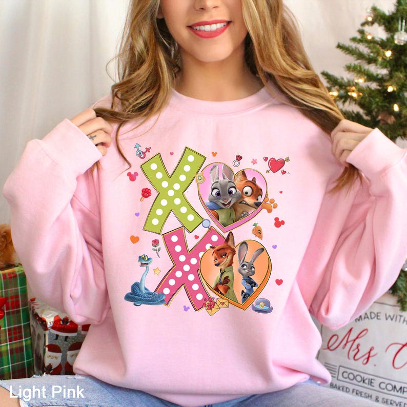 May include: Light pink sweatshirt featuring the characters Judy Hopps and Nick Wilde from Zootopia. The design includes the letters "XO" with heart and character graphics. The text "Light Pink" is at the bottom of the image.