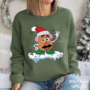 Puede incluir: Sudadera verde militar con un personaje de dibujos animados de Mr. Potato Head con un gorro de Pap&aacute; Noel y luces navide&ntilde;as. El dise&ntilde;o incluye acebo y copos de nieve. La sudadera es de cuello redondo.