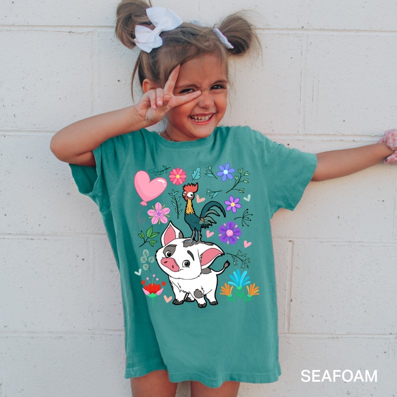 K&ouml;nnte beinhalten: Ein t&uuml;rkisfarbenes T-Shirt mit einem Cartoon-Motiv aus einem Schwein und einem Hahn. Das Schwein ist wei&szlig; mit schwarzen Flecken und der Hahn ist rot, gelb und schwarz. Das Motiv ist von rosa, lila und blauen Blumen umgeben.